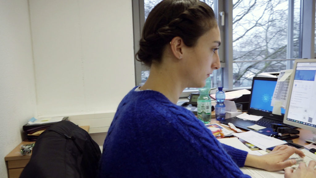 Brünette Kommissaranwärterin sitzt in blauem Pullover am Rechner.
