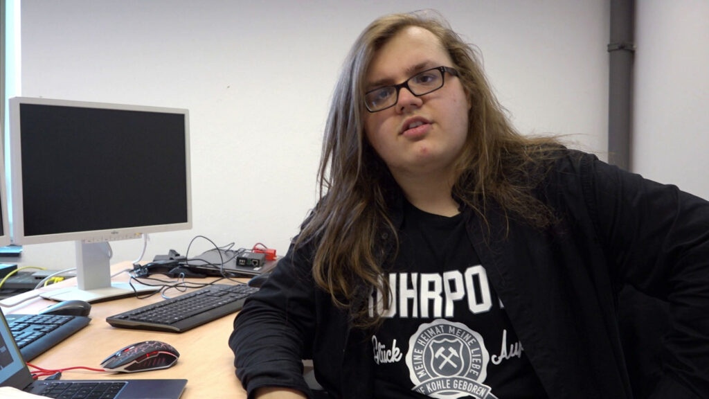 18-Jähriger mit Brille und langen Haaren sitzt an einem Schreibtisch mit Computer und schaut in die Kamera.