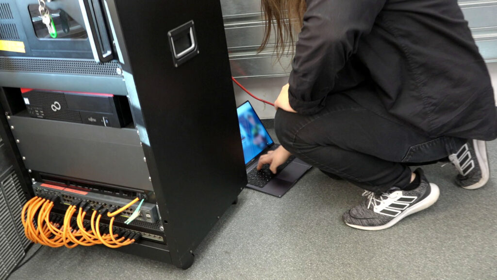 Informatiker der Polizei NRW kniet mit einem Laptop neben einem Rechner.