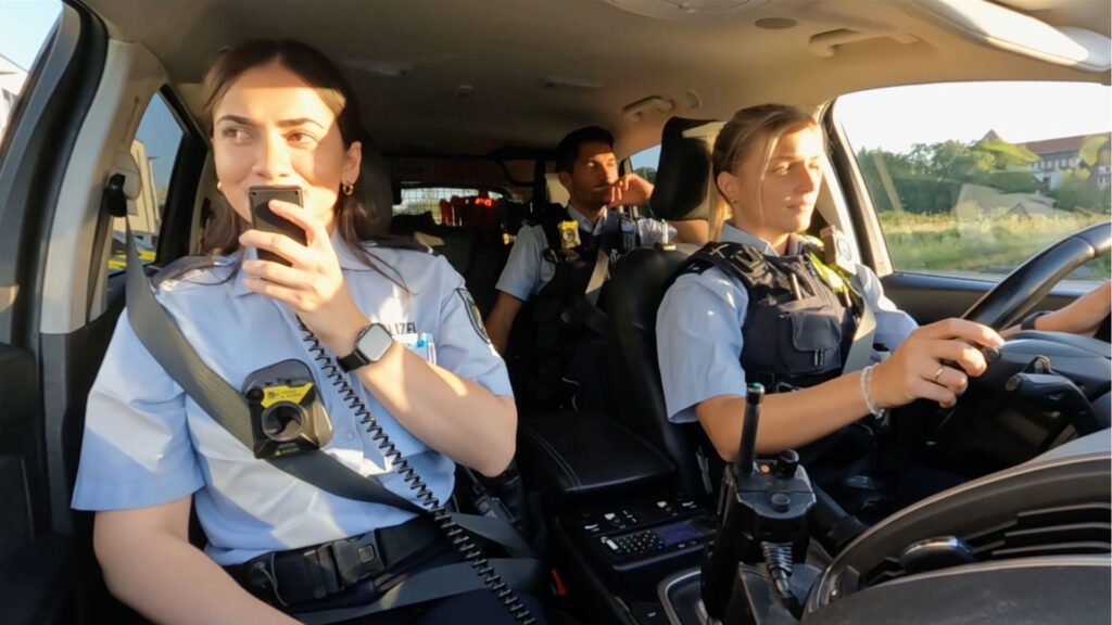 Blick in den Streifenwagen während der Fahrt aus Perspektive der Frontkonsole. Auf dem Beifahrersitz funkt eine dunkelhaarige Kommissarsanwärterin, während auf der Fahrerseite eine blonde Polizistin hinterm Steuer sitzt. Beide tragen ihre Haare zu einem Zopf zusammen gebunden. Auf der Rückbank hinter der Fahrerin sitzt ein Polizist mit dunkelbraunen Haaren. Alle Fahrzeuginsassen tragen Bodycams und Kurzarmhemden der Polizeiuniform. Die Polizisten auf der Fahrerseiten tragen zudem Außentragehüllen.