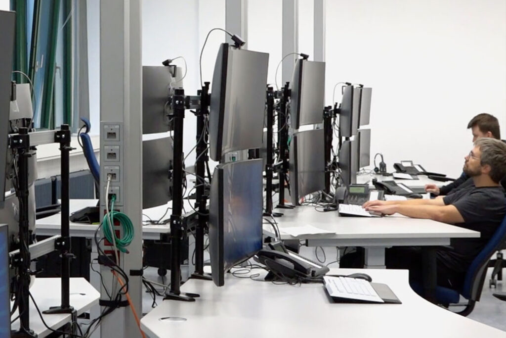 Ein Raum, der zu Schulungszwecken dient und eine Leitstelle simulieren soll. Mehrere Schreibtische stehen sich in Reihen gegenüber. Auf jedem Schreibtisch sind zwei curved Computerbildschirme übereinander angebracht, wobei der obere Bildschirm ebenfalls mit einer Kamera ausgestattet ist. Zudem steht auf jedem Schreibtisch ein Telefon, eine Computermaus sowie eine Computertastatur. Im hinteren Teil des Raum sind zwei Schreibtische mit zwei arbeitenden Männern besetzt.